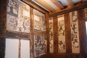 Religious texts and imagery could be found everywhere in the interiors of middling houses. These visuals displayed not only belief but status and wealth. These wall paintings are in a first-floor room of what is now the town council offices in Ledbury, Herefordshire. The property was originally a house of middling status built in the early 1500s and extended in about 1600. The wall paintings probably date from between 1575 and 1600 when the town constable lived there.<br />
<br />
The lower section of the painting mimics wood panelling, which was a popular yet costly form of decoration at this time. The middle of the wall is filled with a repeated pattern of overlapping shapes containing colourful flowers, set onto a dark background. Towards the top of the wall, just below the ceiling, is a painted frieze with biblical texts.<br />
<br />
These selected passages have a strong moral tone, emphasising the benefits of following Gods commandments and being a good citizen. Such decoration reminded the viewer of their moral and religious responsibilities, but it also displayed the homeowners piety as well as their role as a civic leader. These were both important elements of urban middling status.