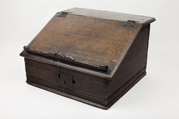 This sloping desk box might feel familiar to those using laptop stands or writing mats today.  It was used to prop up books and documents for reading or writing. It was also used for storing writing materials, from documents and paper to ink. The two hinges seen on the top of the box allow the lid to be lifted, giving access to the items stored within. Similar to musical instruments like the virginals, it could be moved around the room, stored, and then placed on a table for use. Desks, as we know them today, did not become popular until the late seventeenth century, so many middling households owned writing boxes like this. If you saw one in a household, it was a clear sign that the occupier was literate, perhaps involved in business or trade or held a role in the community, like churchwarden, that required literacy. 